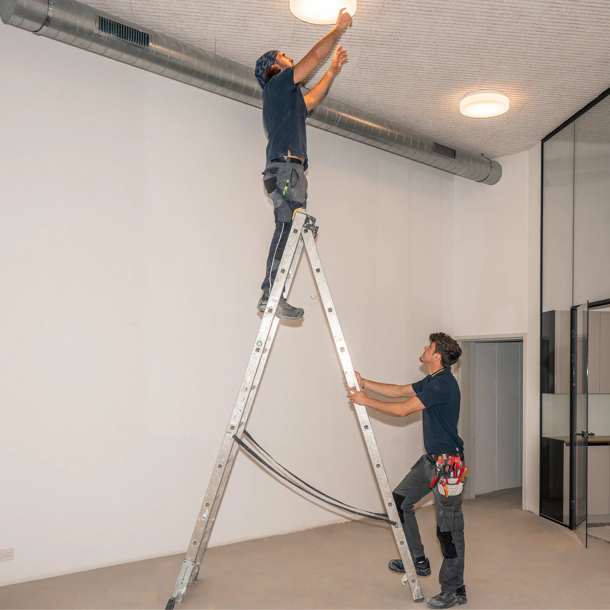Zwei Elektriker der elektro scherzinger ag montieren LED-Deckenleuchten von der Leiter aus